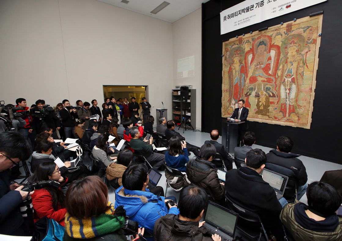 Um representante da CHA está na frente da pintura da Tríade Shakyamuni, o primeiro item devolvido à Coreia em 2014 como parte deste projeto.